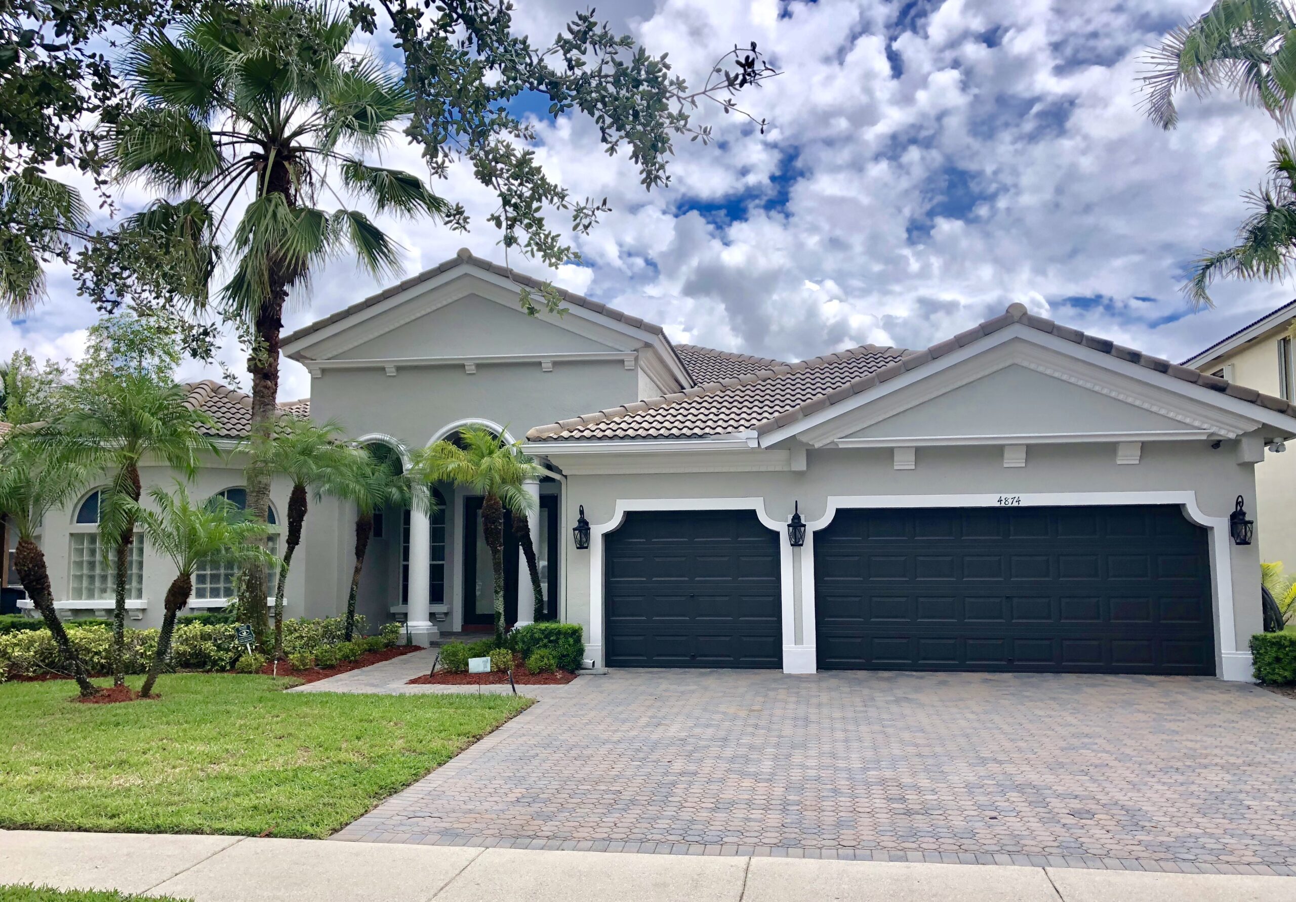 A modern single-story home with a tiled roof, three car garage, and lush landscaping under a partly cloudy sky.